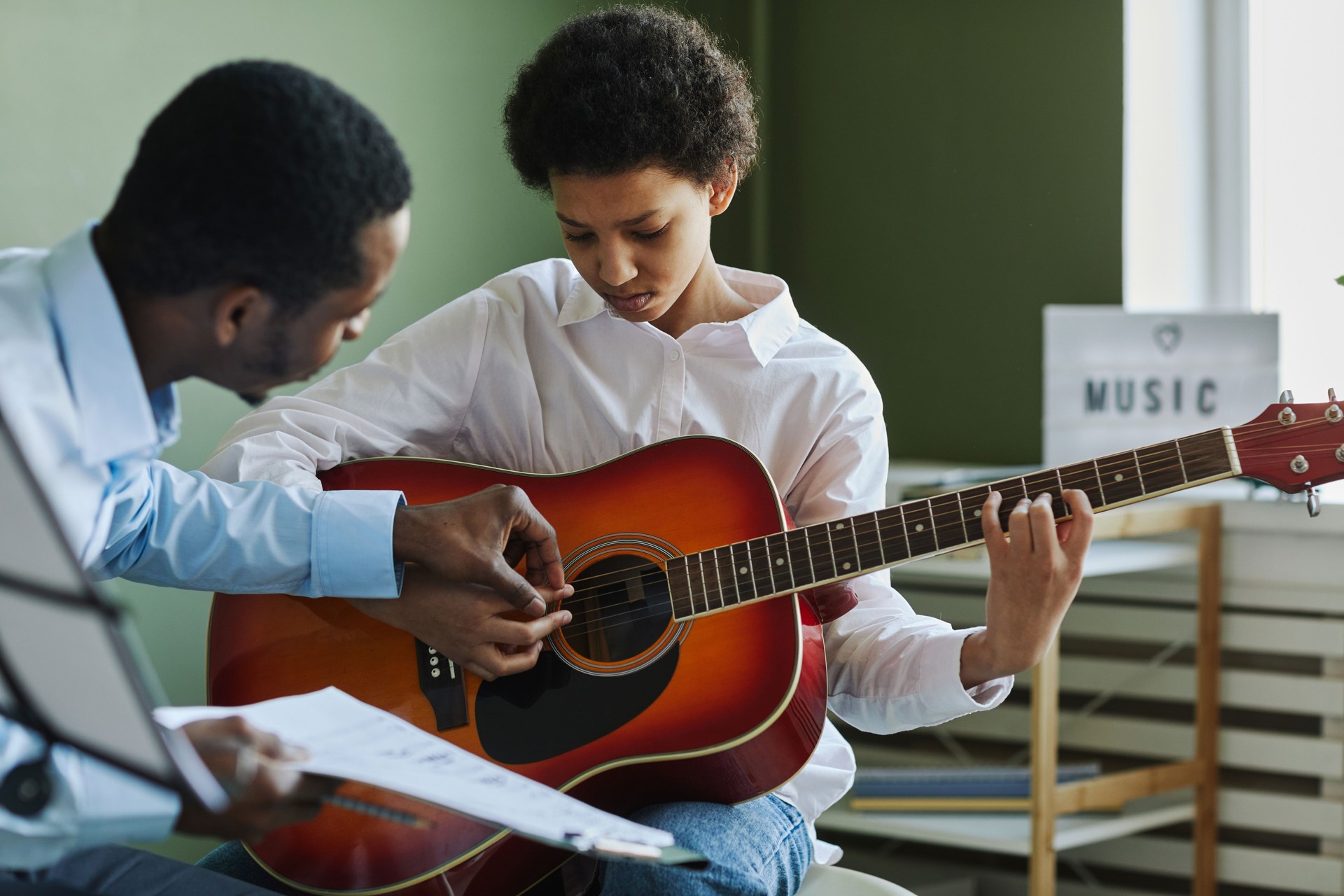 Youthful diligent girl playing acoustic guitar while teacher consulting her