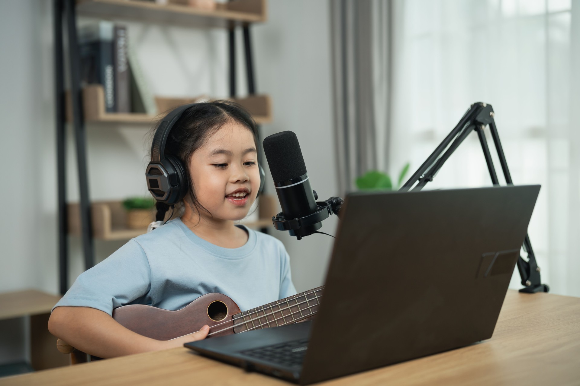 Young girl with headphones sings and plays ukulele in front of laptop during online music lesson in modern home setup with natural light.